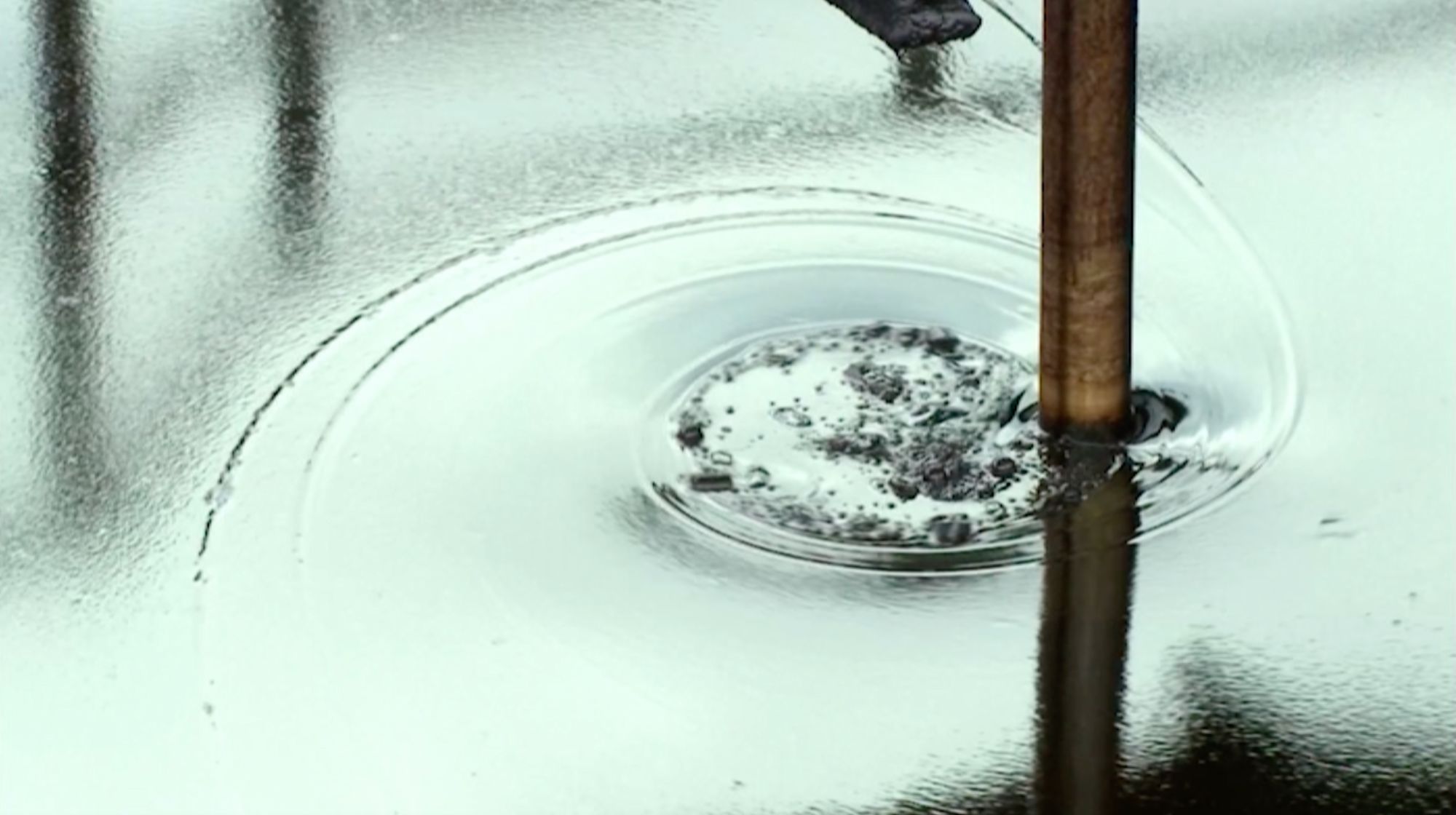 Nahaufnahme: Ein Metallstab taucht in trübes Wasser mit dunklen Schwebeteilchen, Wellenkreise auf der Oberfläche