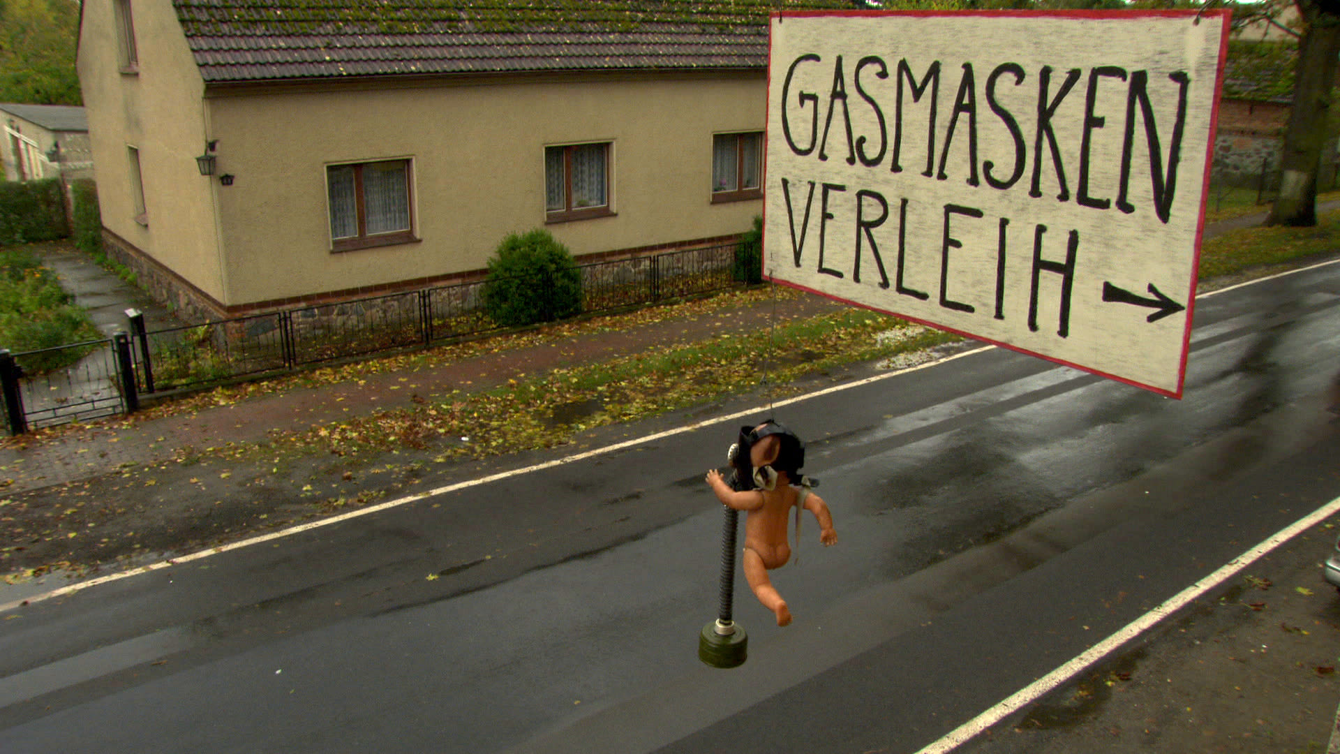 Baby-Puppe mit Gasmaske an einem Pfosten vor einem Wohnhaus, daneben Schild „Gasmasken-Verleih“