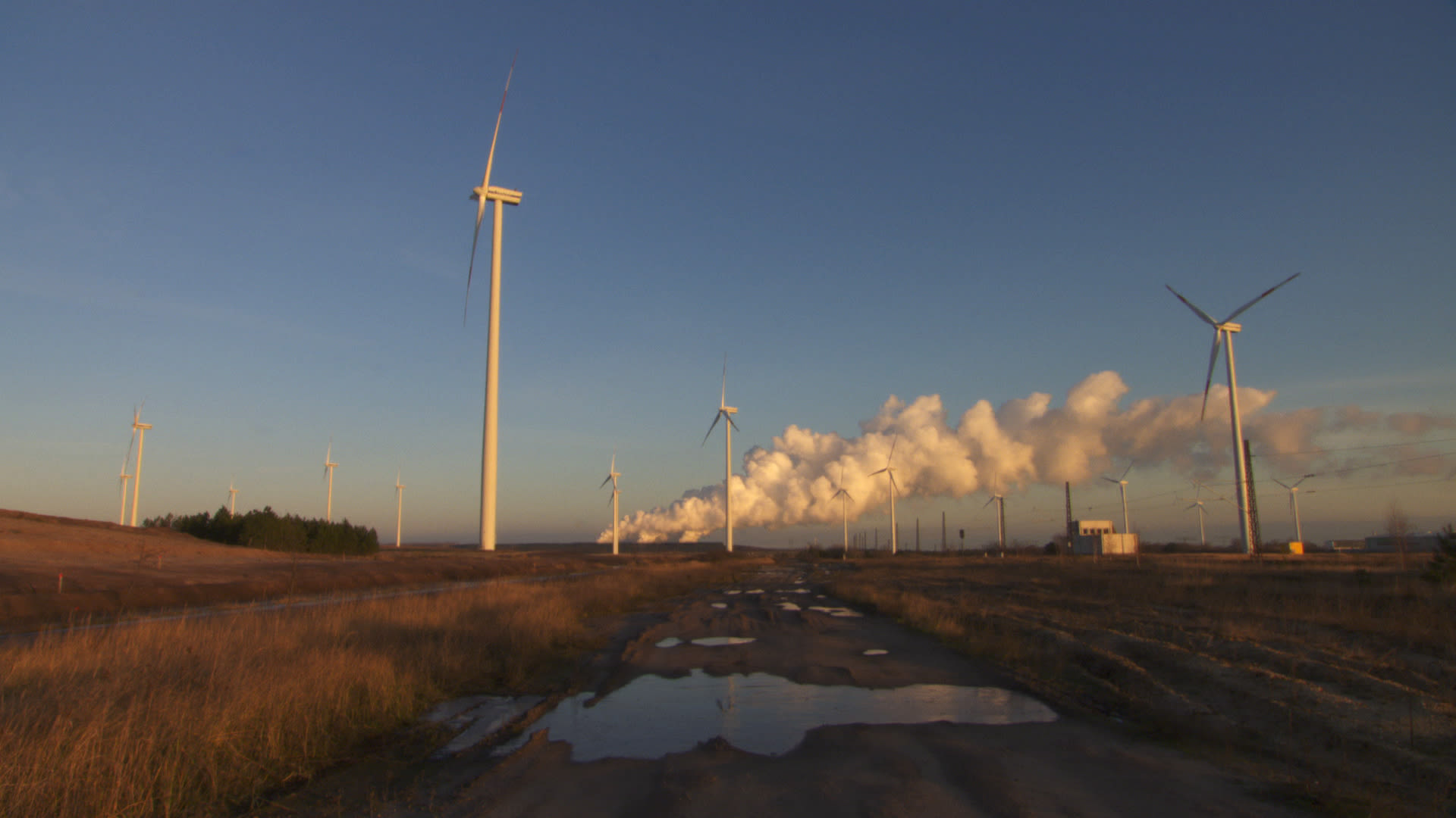 Windräder bei Abenddämmerung vor aufsteigenden Dampfschwaden eines Kraftwerks, Pfütze im Vordergrund
