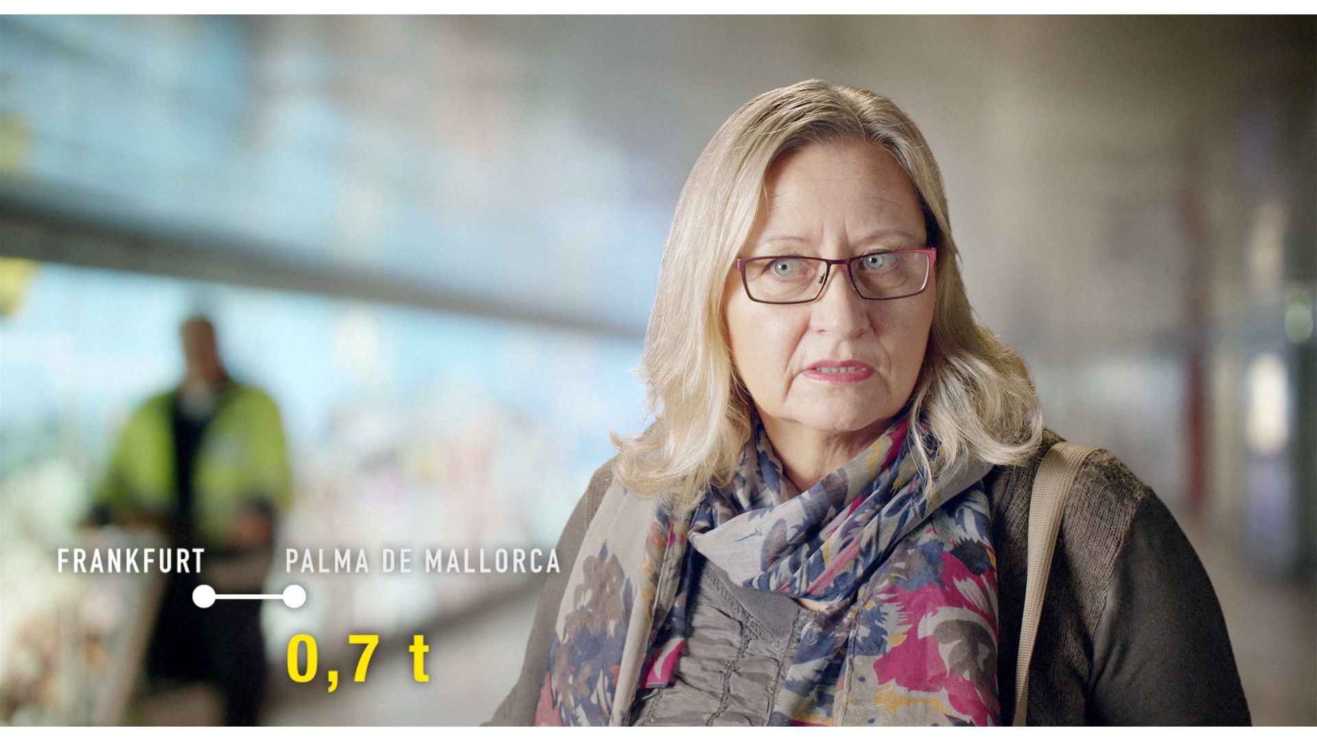 Frau mit Brille und buntem Schal am Bahnhof, Einblendung „Frankfurt – Palma de Mallorca, 0,7 t“