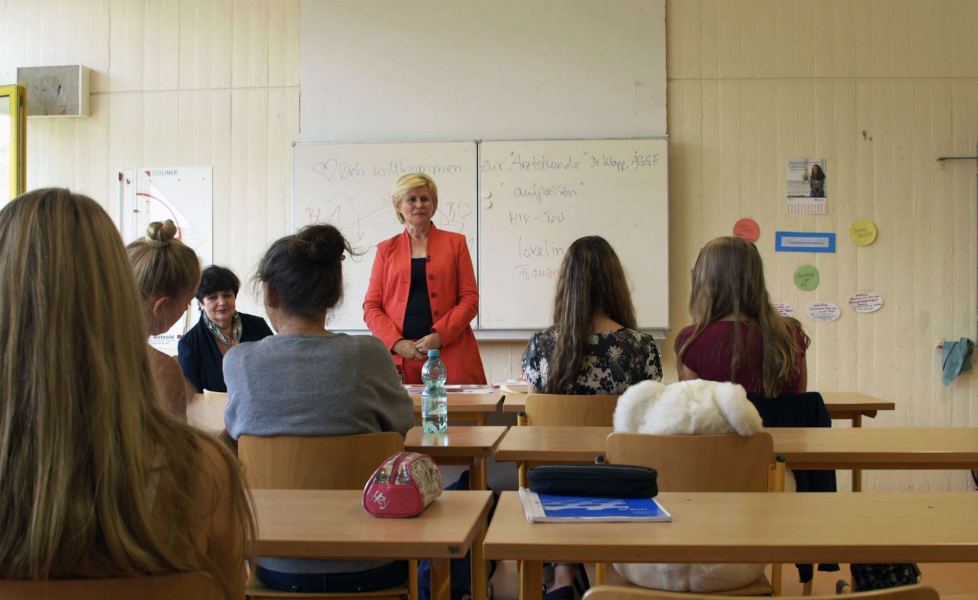 Frau im roten Blazer spricht vor einer Schulklasse, Tafel zeigt „Herzlich willkommen zur Arztstunde“