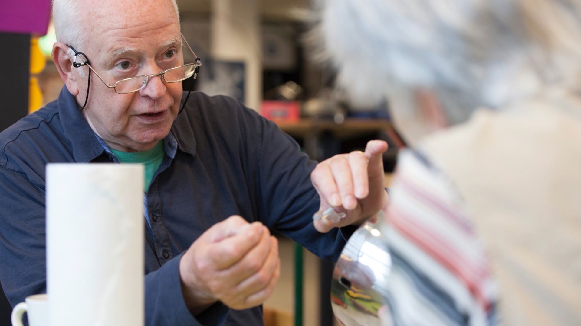 Ein älterer Mann mit Brille gestikuliert im Gespräch in einem Werkstattraum
