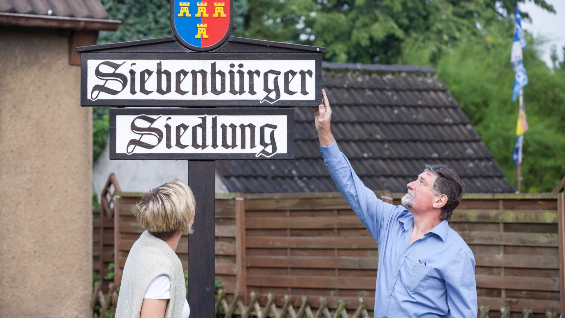 Ein Mann zeigt einer Frau auf ein Ortsschild mit der Aufschrift „Siebenbürger Siedlung“