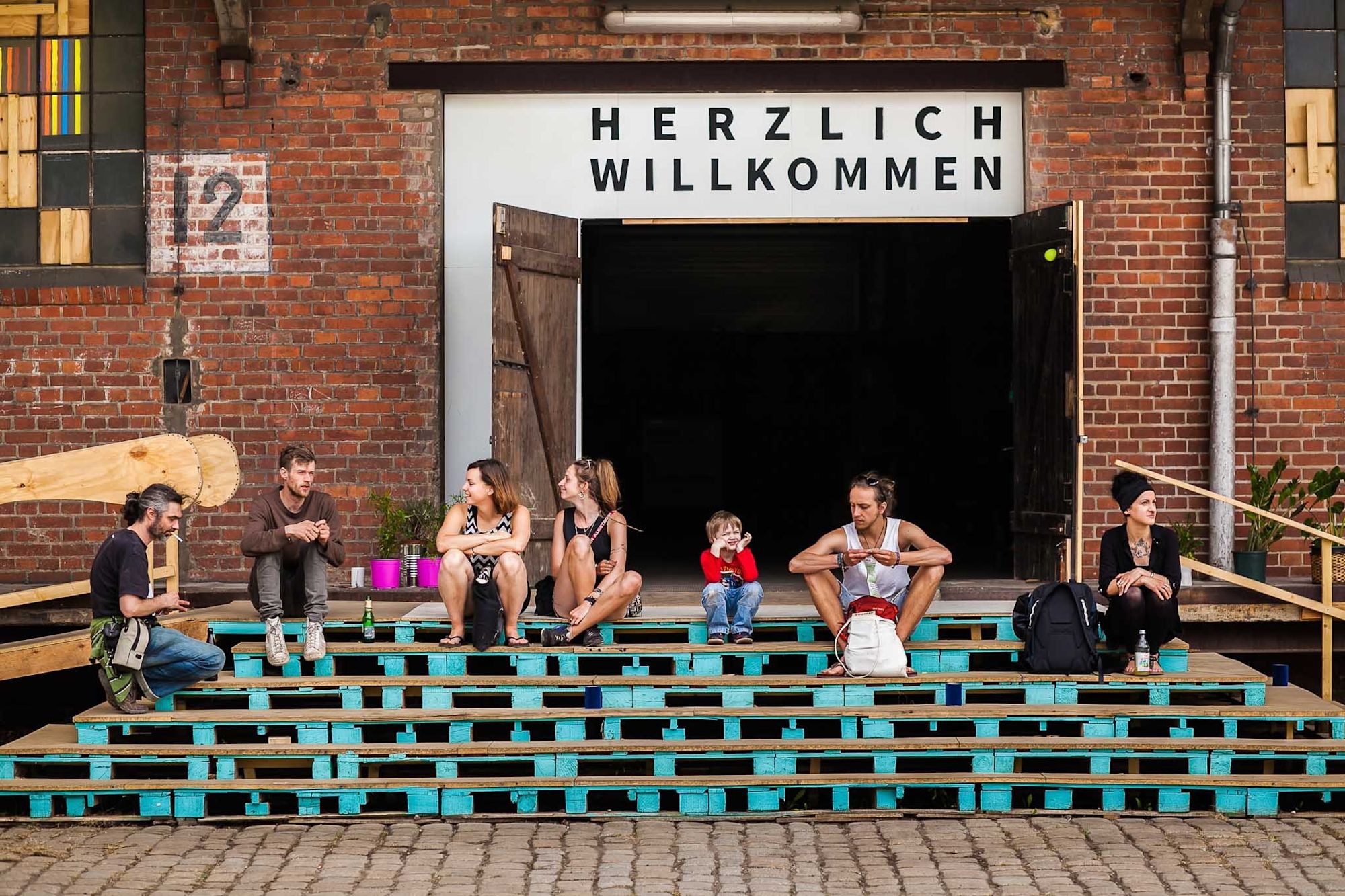 Menschen sitzen auf türkisen Palettenstufen vor Tor mit Schriftzug „Herzlich Willkommen“