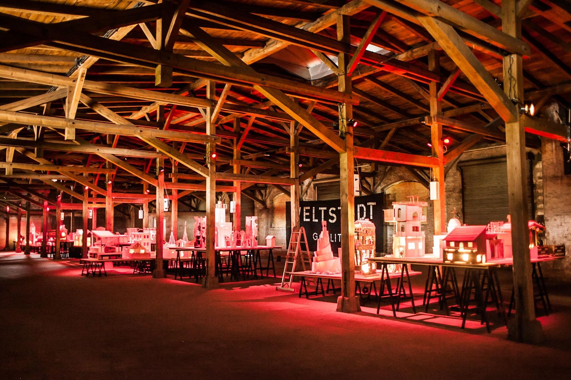 Rot beleuchtete Lagerhalle mit Holzdachstuhl und Pappmodell-Stadt, Schriftzug „Weltstadt“