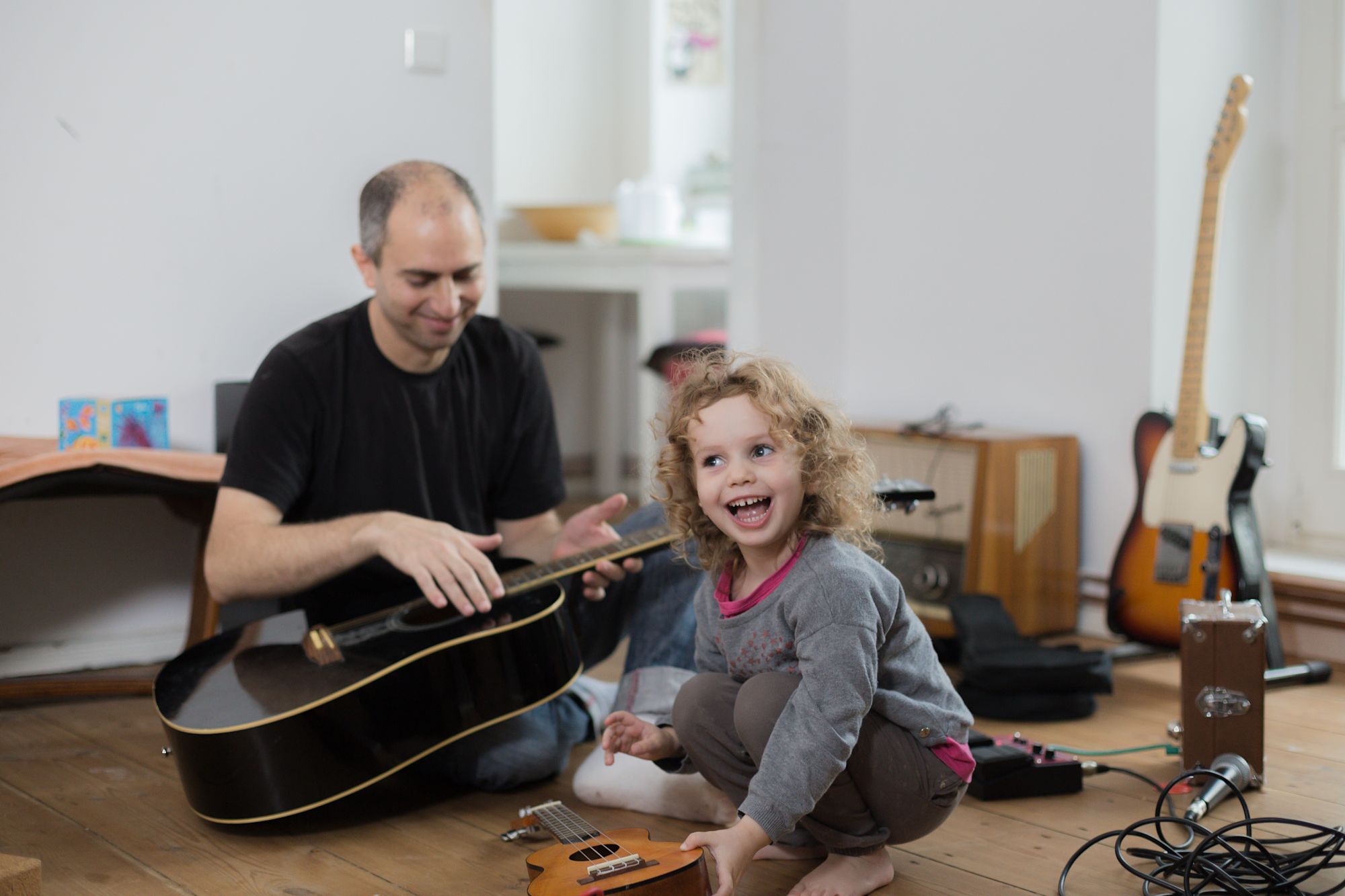 Vater spielt Gitarre, lachendes lockiges Mädchen sitzt vor einer Ukulele