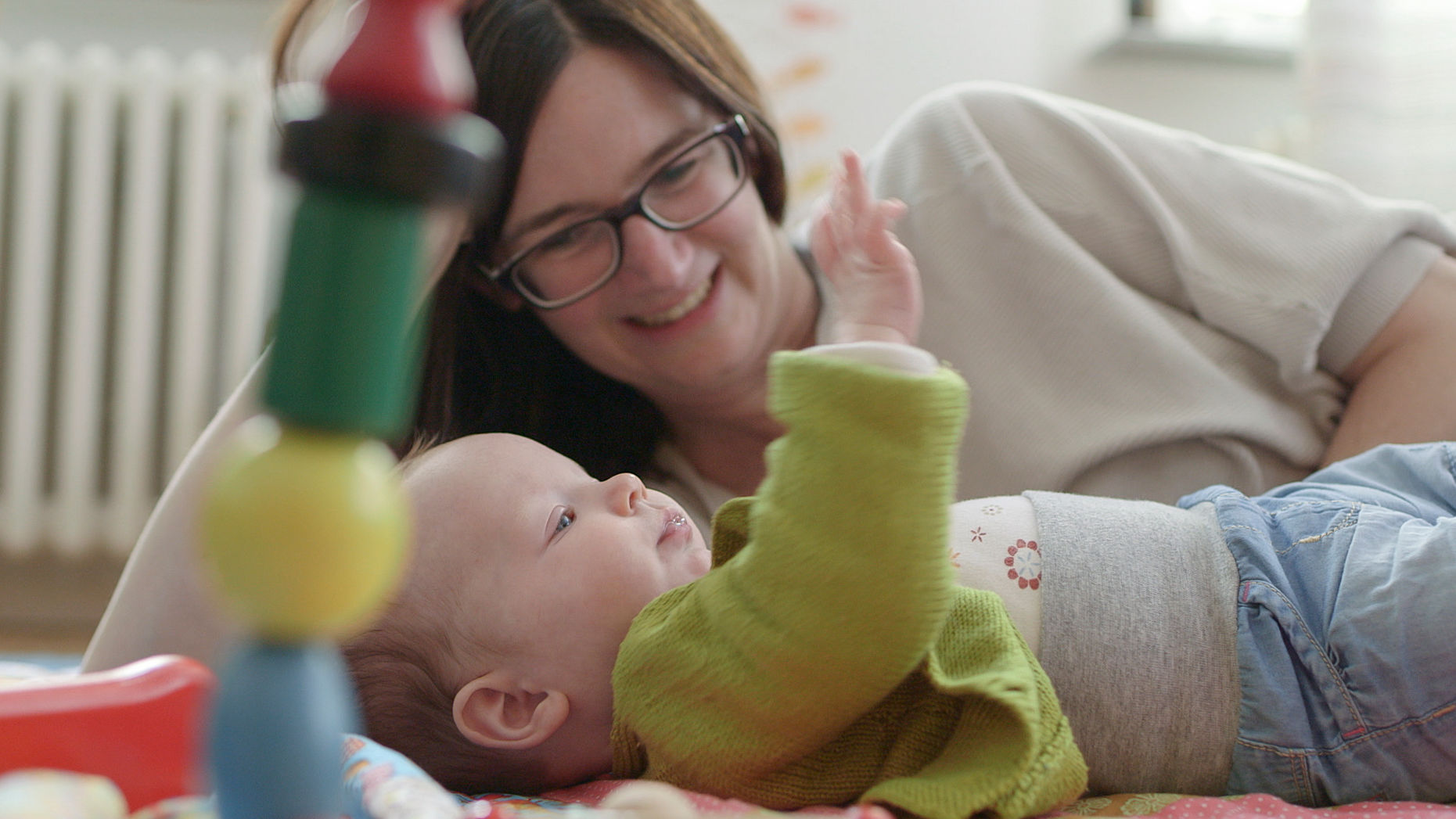 Mutter liegt neben Baby auf der Krabbeldecke, buntes Rasselspielzeug im Vordergrund