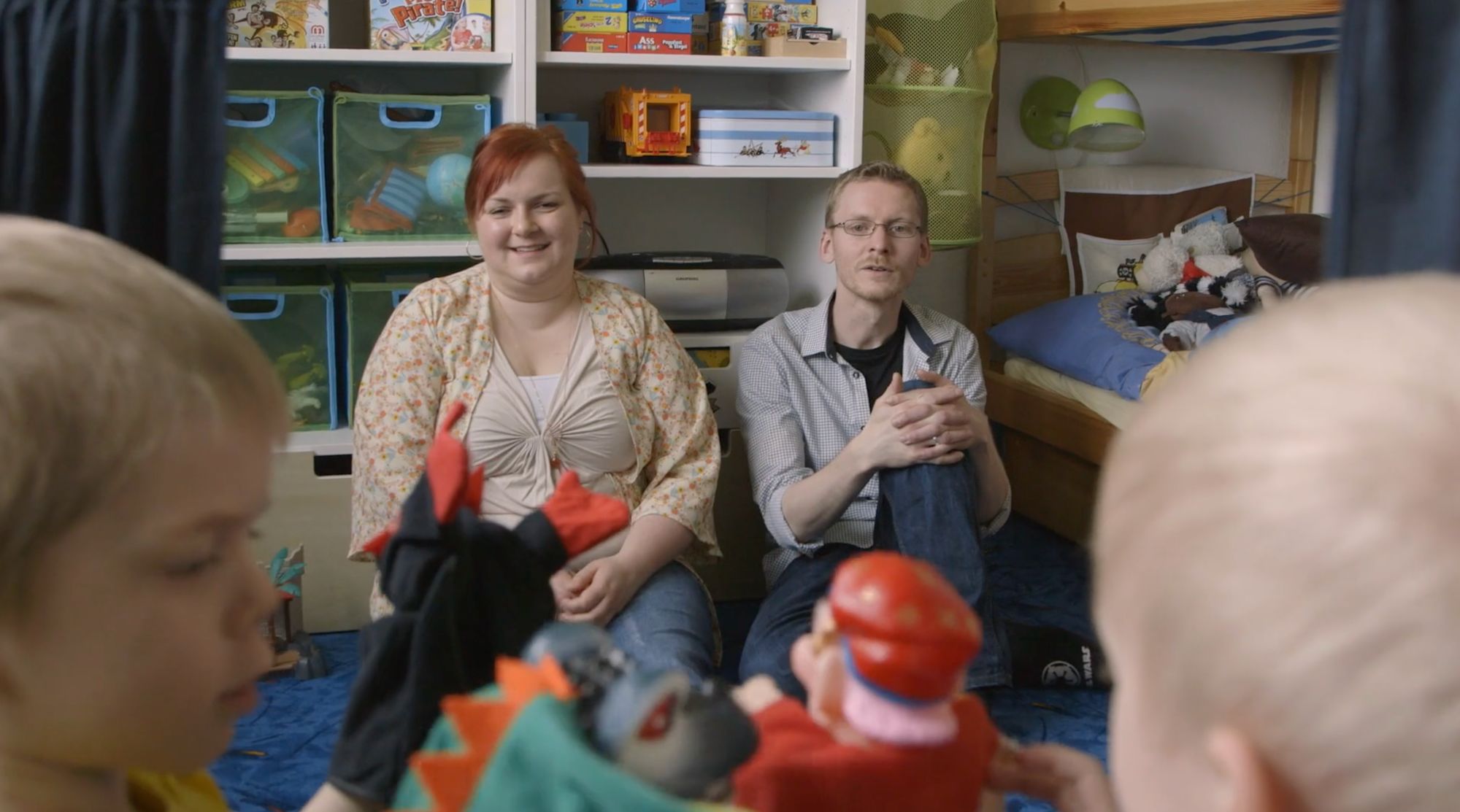 Paar sitzt im Kinderzimmer auf dem Boden, im Vordergrund Handpuppen der Kinder