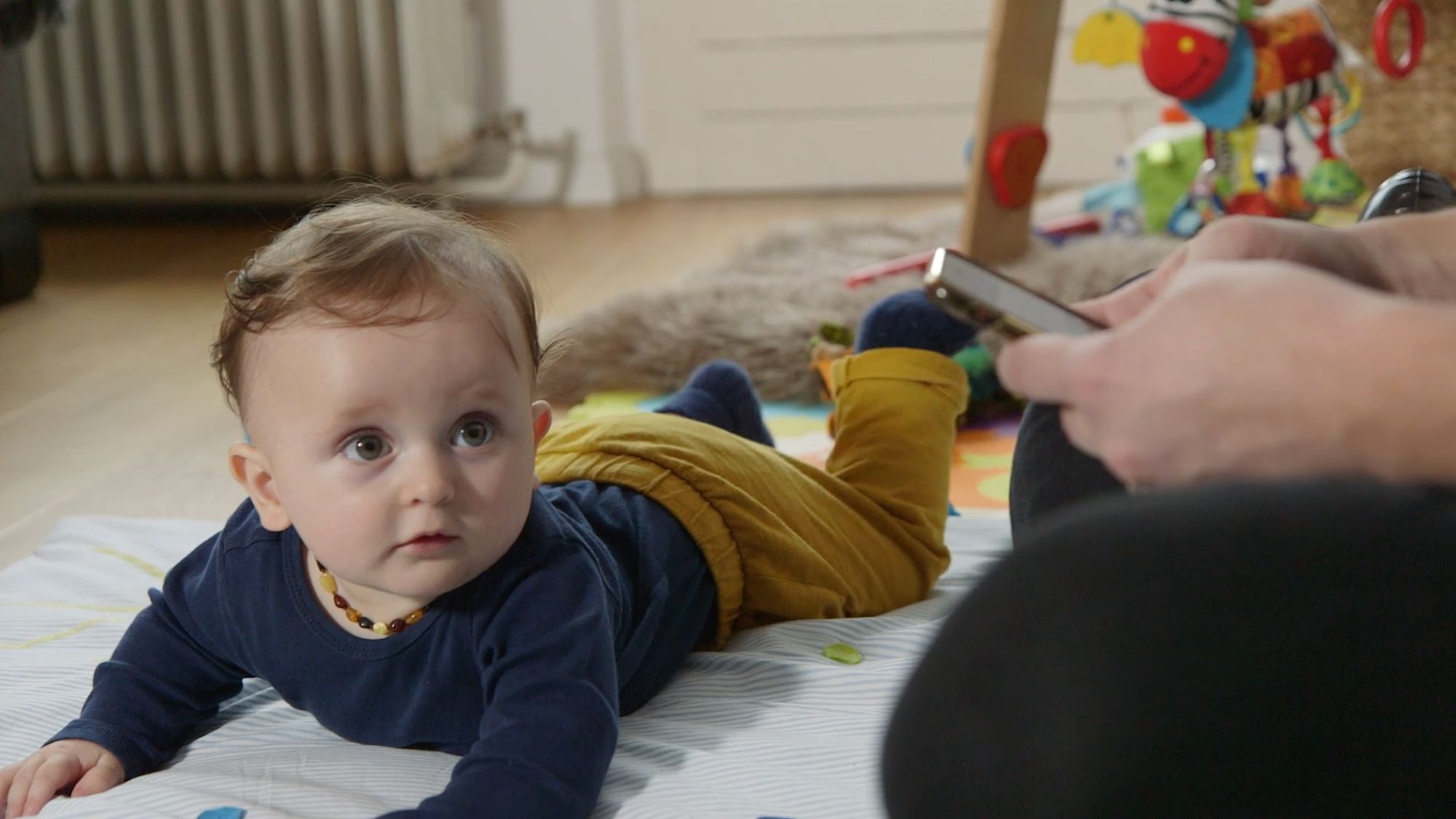Baby stützt sich auf der Krabbeldecke ab, Erwachsenenhand hält im Vordergrund ein Handy