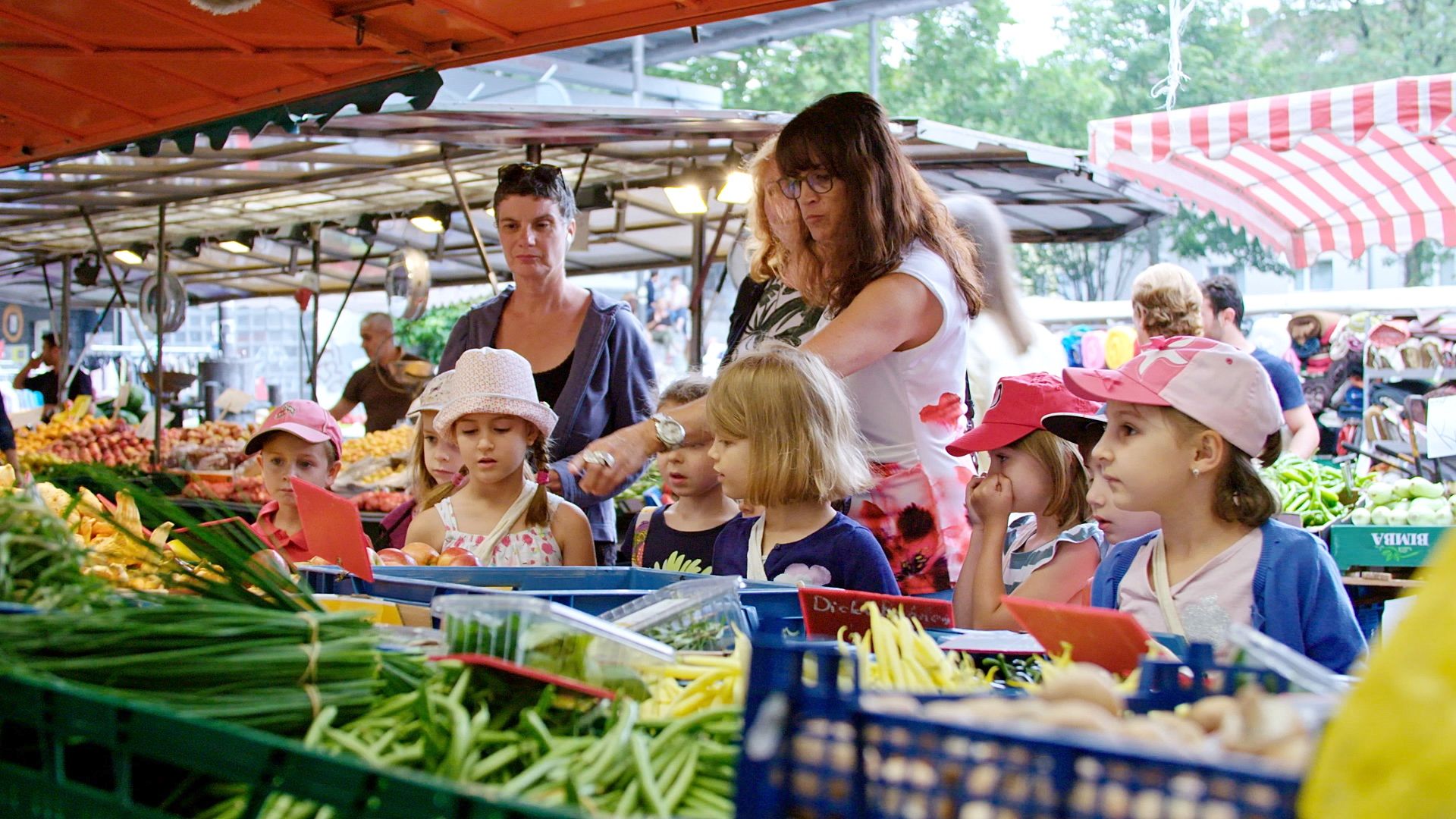 Kinder mit Hüten stehen an einem Marktstand voller Gemüsekisten