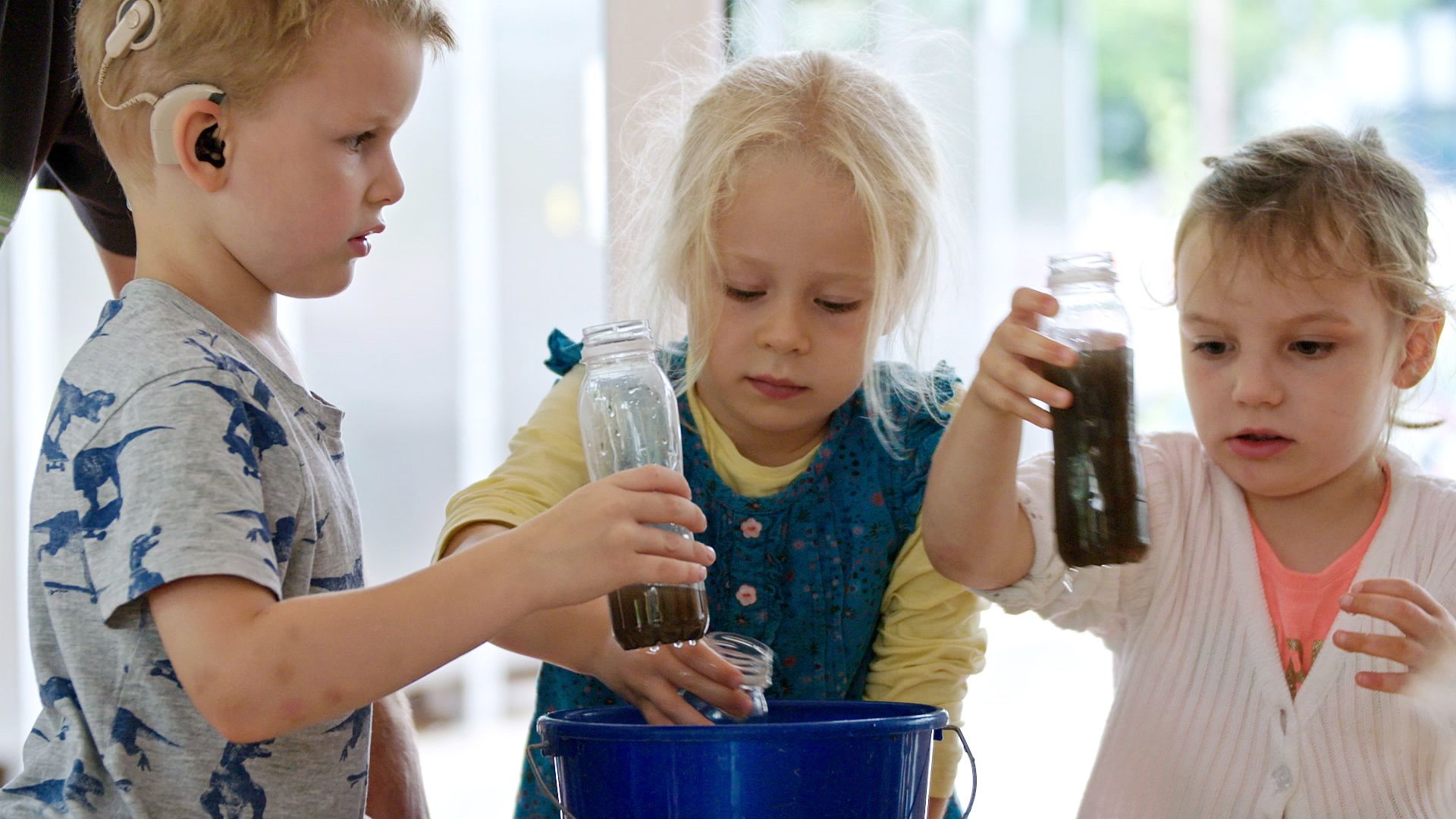 Drei Kinder füllen dunkle Flüssigkeit aus Gläsern in einen Eimer