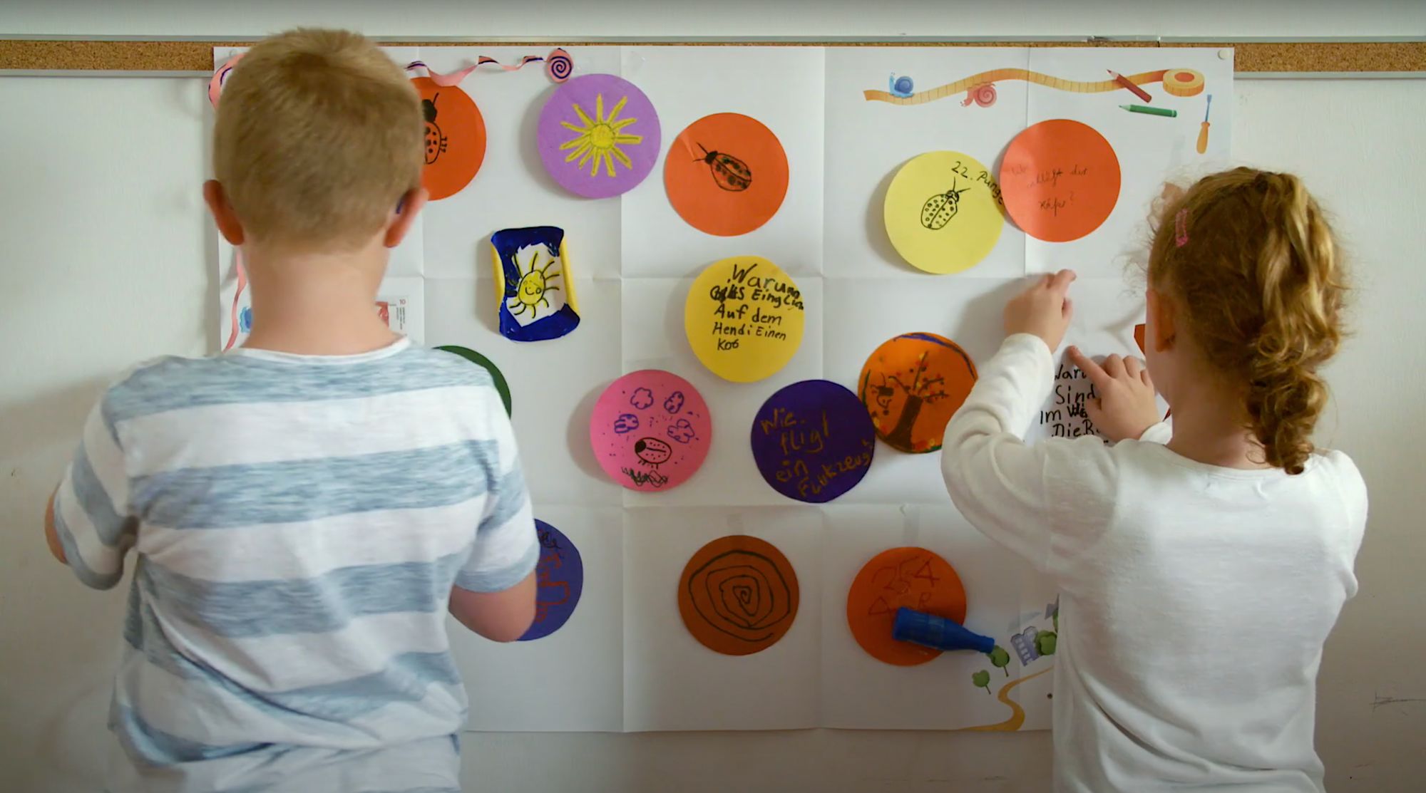 Zwei Kinder befestigen bunte Papierkreise mit Fragen an einer Wand
