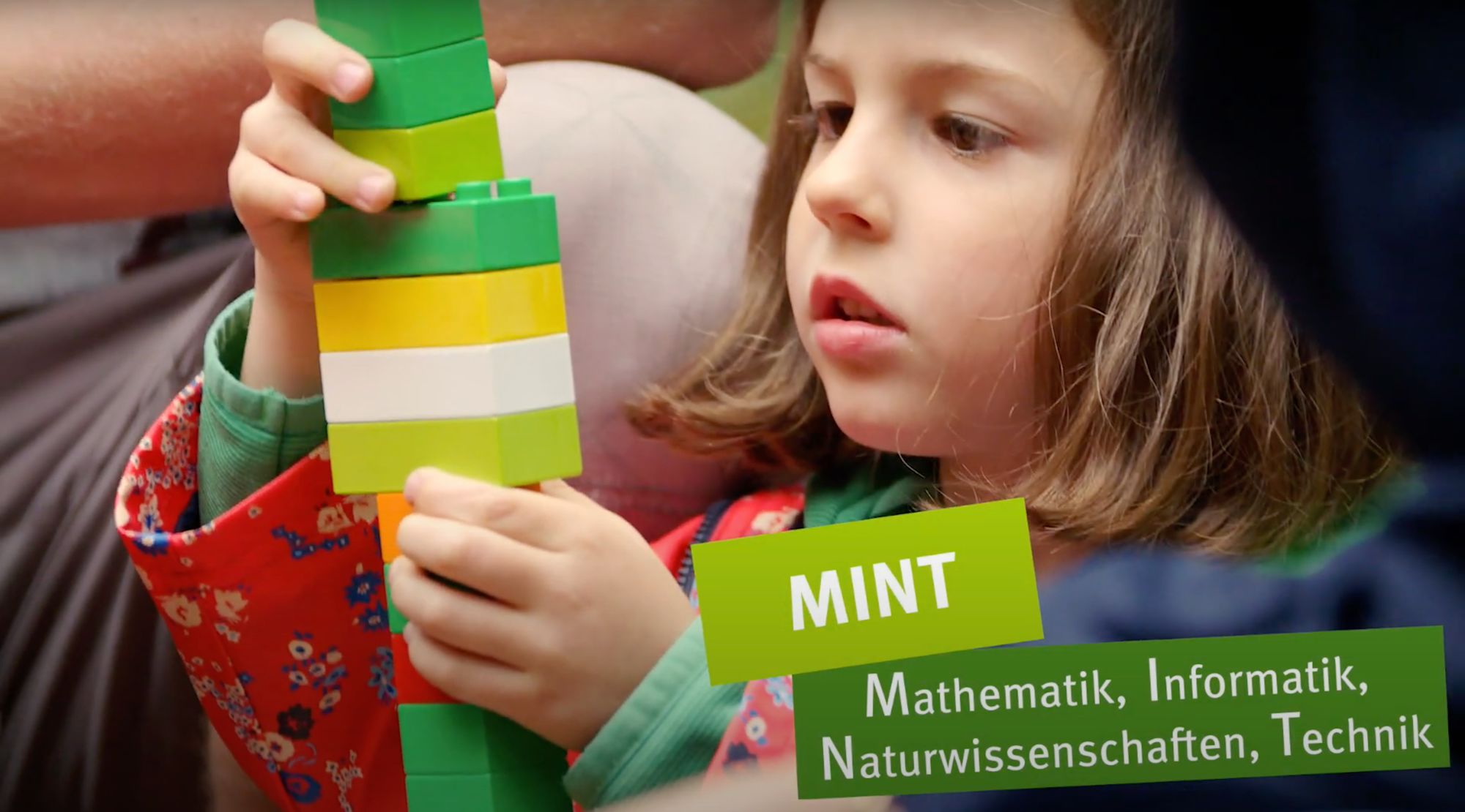 Mädchen stapelt bunte Bausteine, Texttafel nennt den Begriff „MINT“
