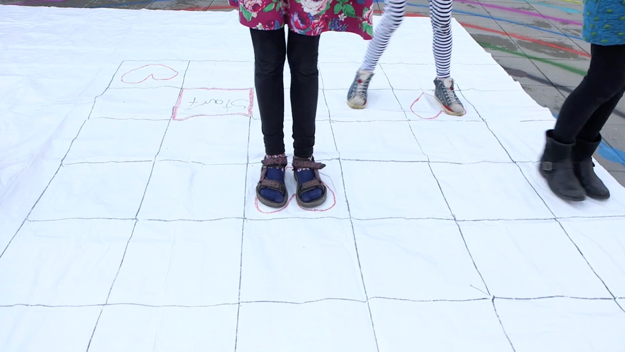 Kinderfüße stehen auf einem großen, bemalten Stoff-Spielfeld mit Startfeld im Freien