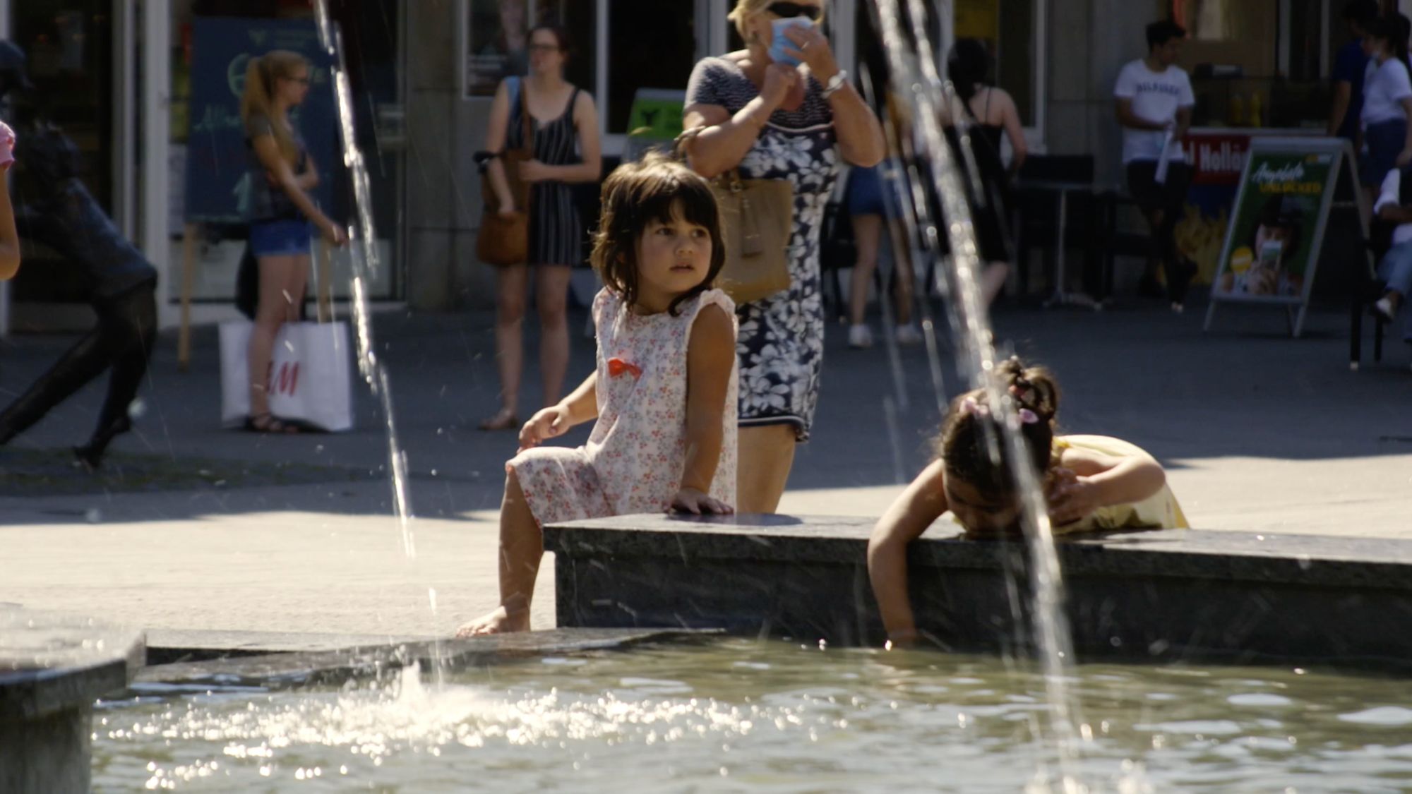 Kinder kühlen sich an einem Brunnen in der Fußgängerzone ab, Passanten dahinter