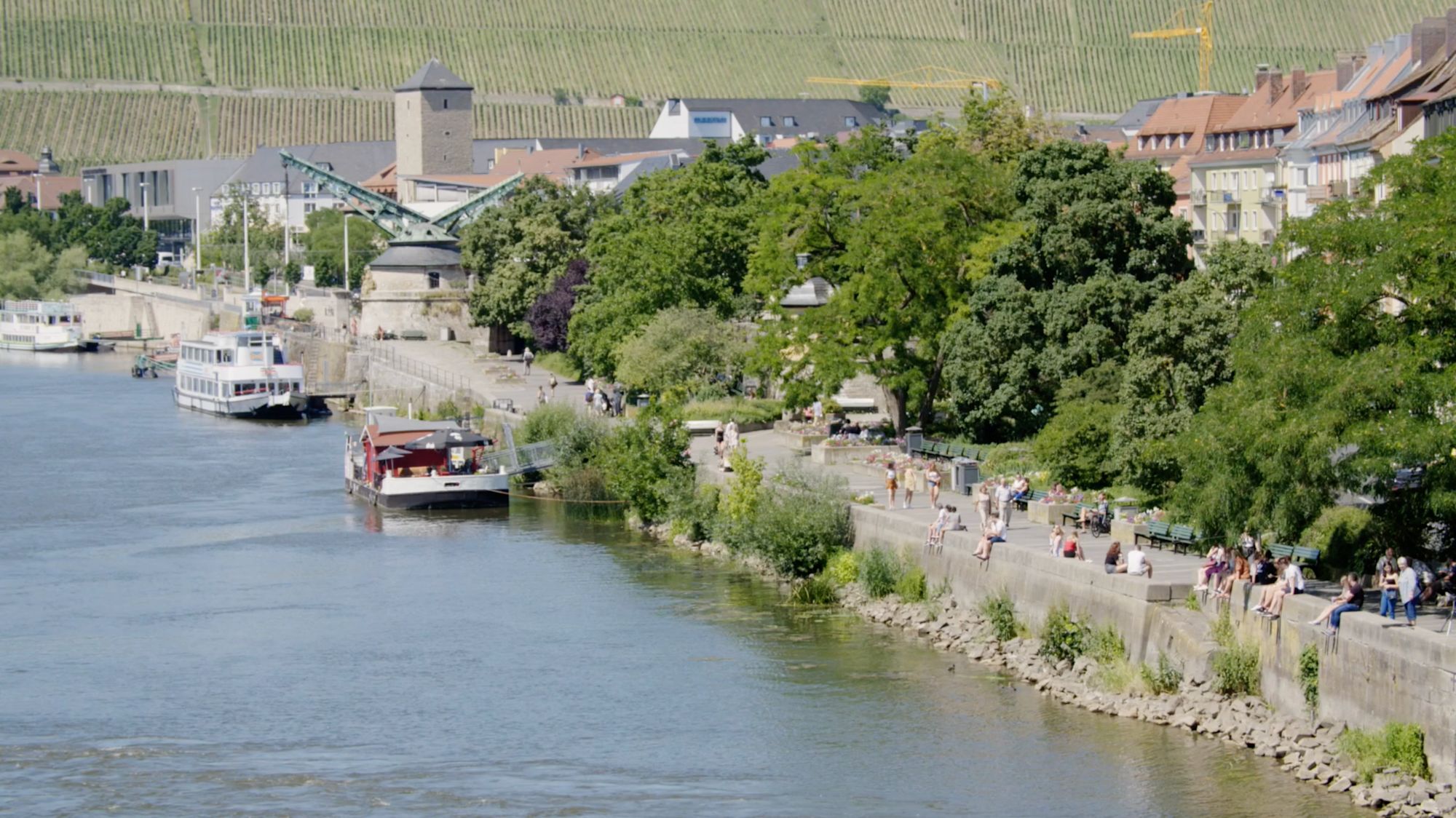 Flusspromenade mit Ausflugsschiffen, Spaziergängern und Weinbergen im Hintergrund