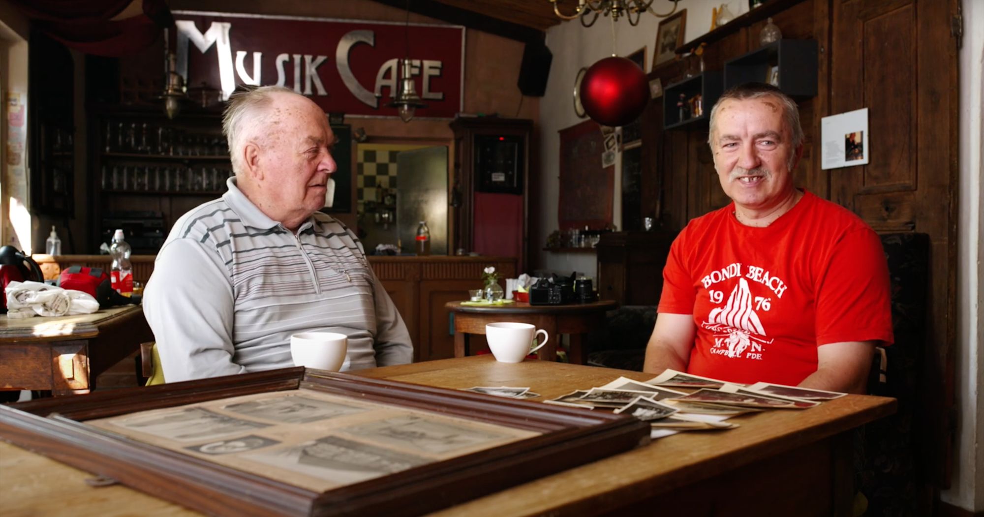 Zwei Männer sitzen im „Musik Café“ und betrachten alte Fotografien auf dem Tisch
