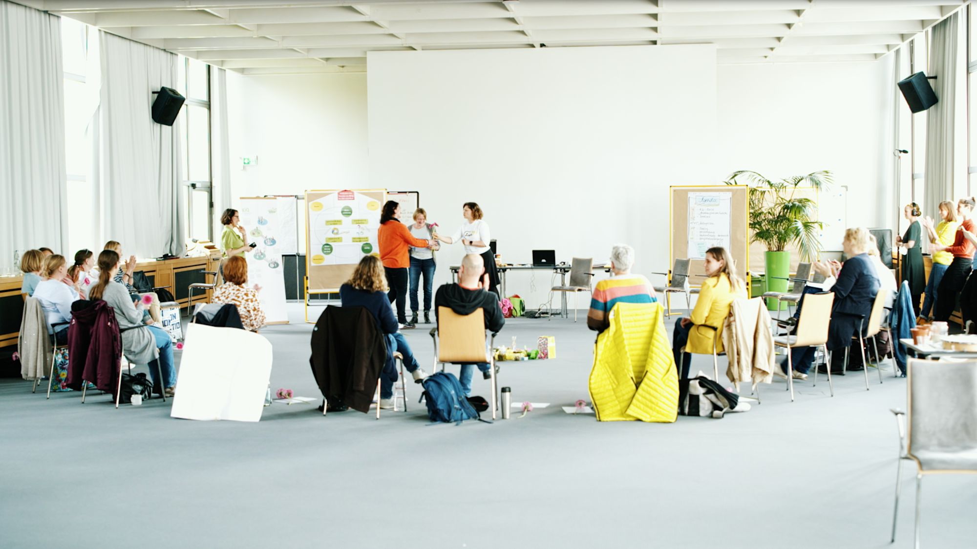 Stuhlkreis in großem Saal, in der Mitte übergeben sich Frauen Blumen vor Flipcharts