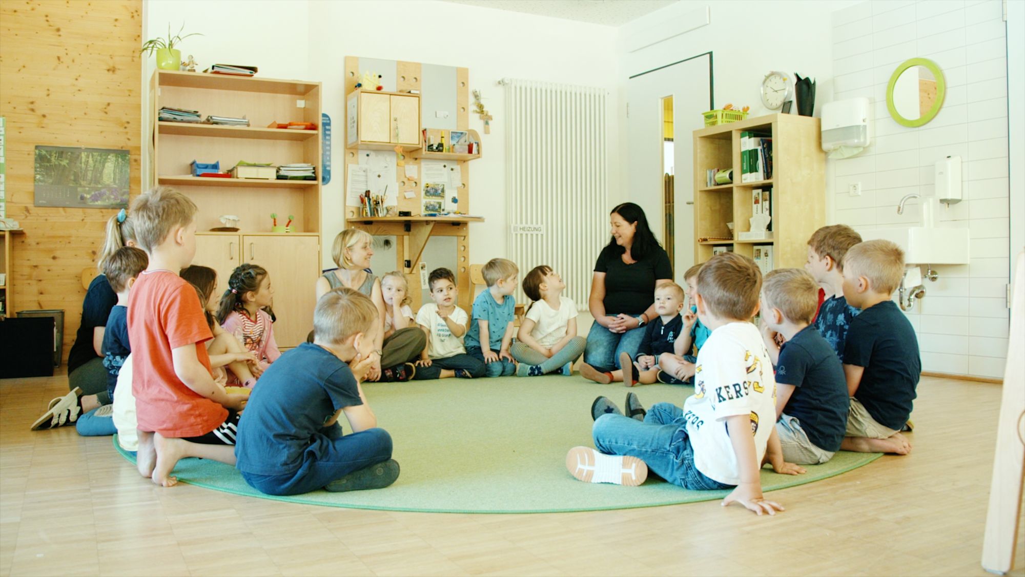 Kinder sitzen im Stuhlkreis auf grünem Teppich, zwei Erzieherinnen in der Mitte