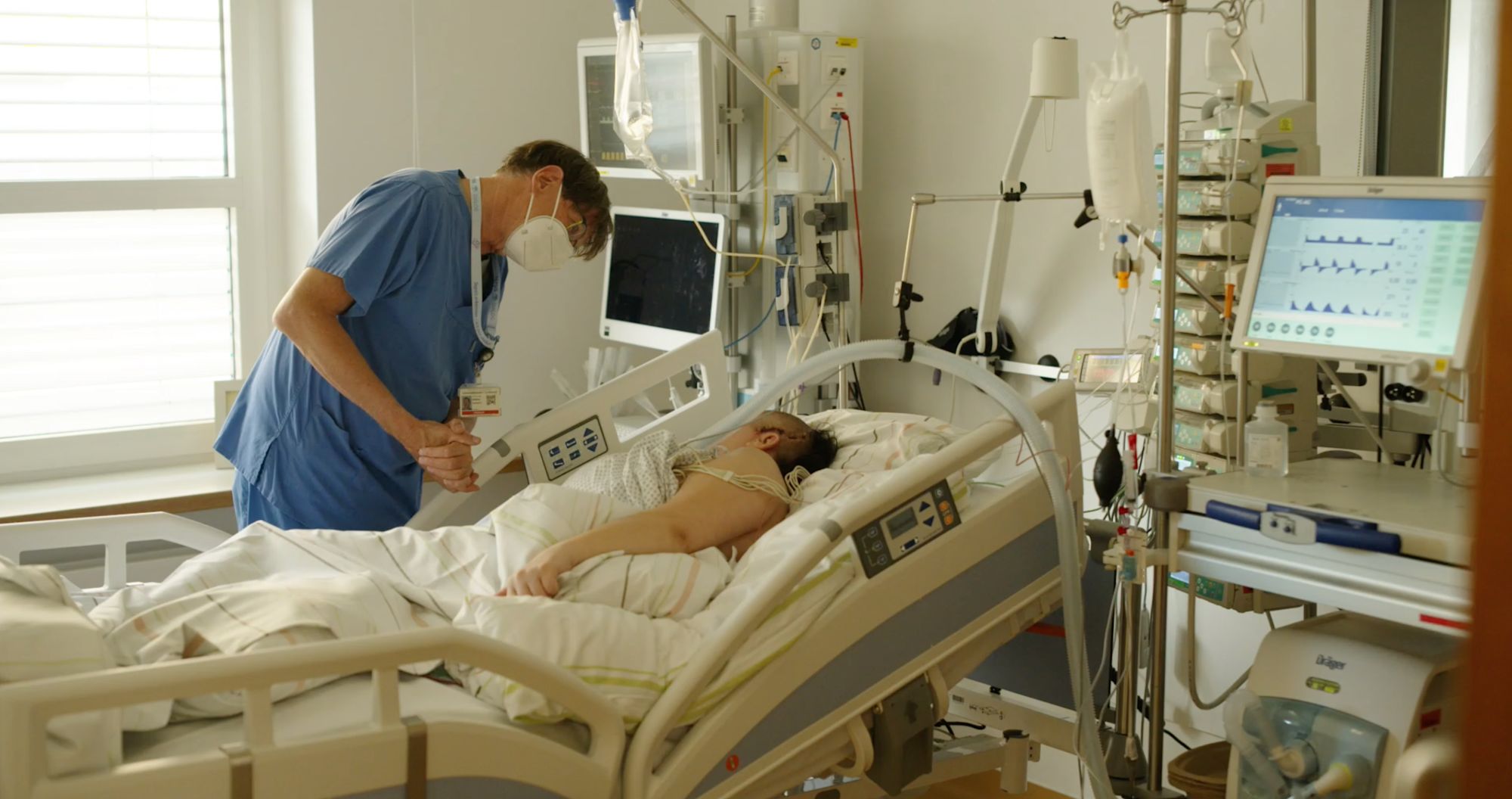 Arzt steht mit gefalteten Händen am Bett eines beatmeten Patienten auf der Intensivstation