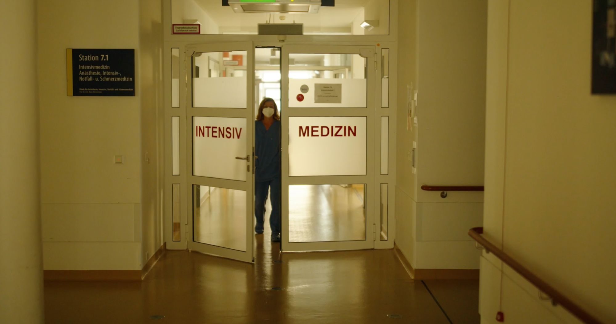 Pflegekraft mit Mundschutz steht hinter der Glastür mit der Aufschrift „Intensiv Medizin“