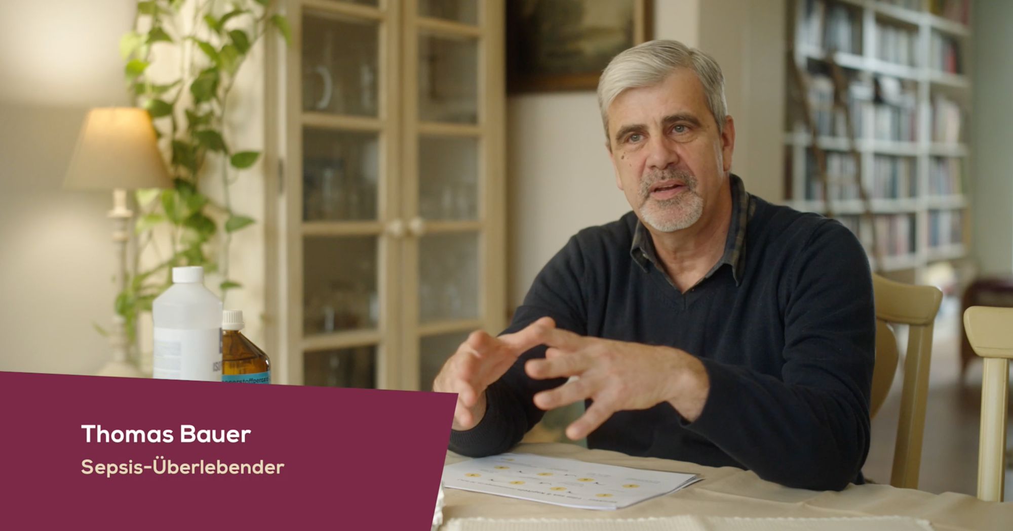 Mann mit grauem Bart gestikuliert am Tisch, Einblendung „Thomas Bauer, Sepsis-Überlebender“