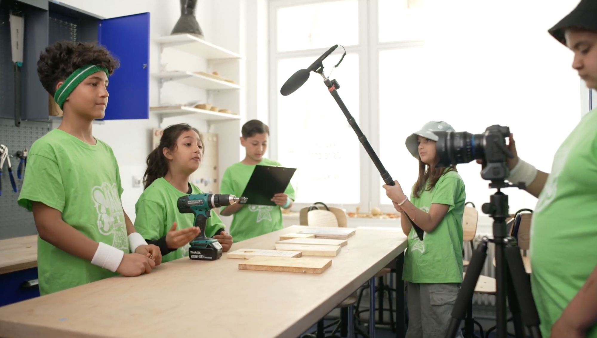 Kinder in grünen Shirts drehen ein Video mit Kamera, Tonangel und Werkzeug in einer Werkstatt