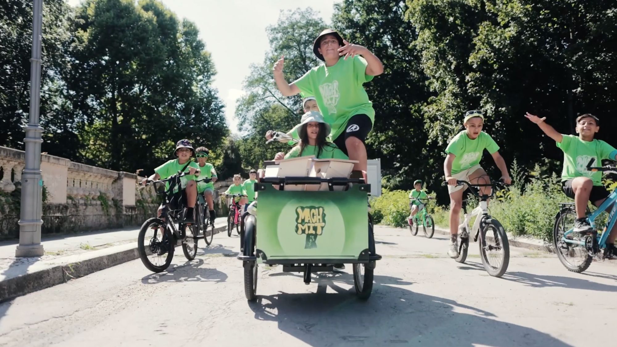 Kinder fahren mit Fahrrädern und einem Lastenrad mit Aufschrift „Mach mit“ durch den Park
