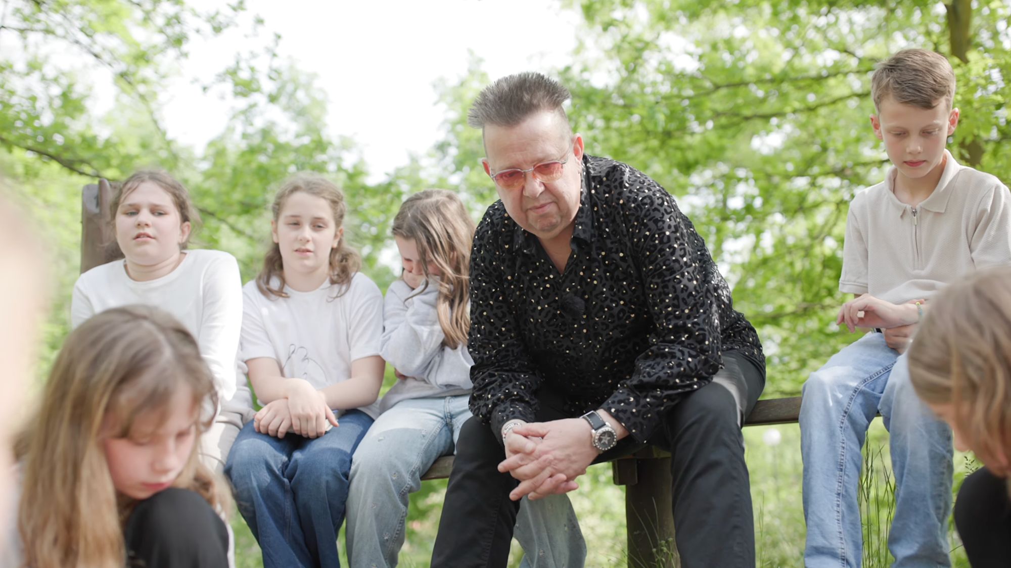 Ein Mann im gemusterten Hemd sitzt mit einer Gruppe Kinder im Wald zusammen