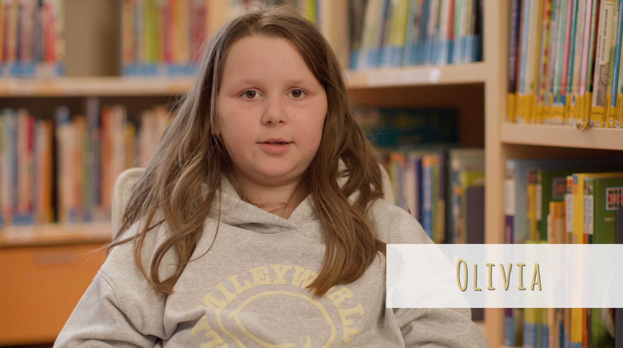 Mädchen mit Pullover spricht vor Bücherregalen, Namenseinblendung „Olivia“