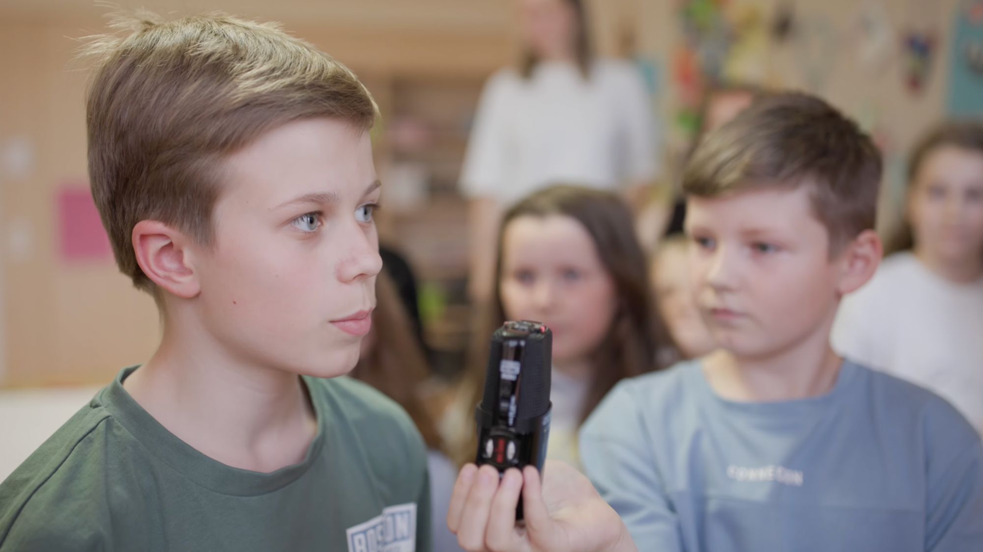 Junge hält ein Aufnahmegerät nah vors Gesicht, im Hintergrund weitere Kinder